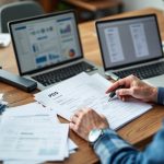Propriétaire assis à un bureau en bois, triant des reçus et des factures scannés sur son ordinateur portable. Piles de papiers et un scanner USB à côté, lumière matinale douce entrant par la fenêtre.