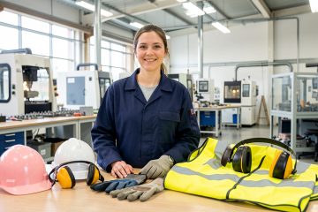 Une responsable sécurité présente différents équipements de protection individuelle disposés sur un établi dans un atelier industriel lumineux.